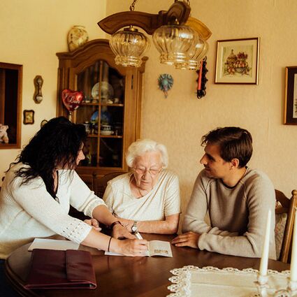 Seniorenbetreuerin sitzt an einem Tisch mit Seniorin und Angehörigem Mann. Sie haben einen Flyer auf dem Tisch liegen.